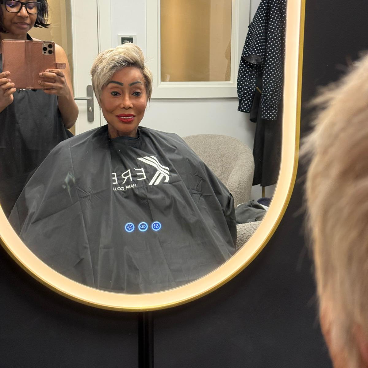 A person with short blonde hair sits in a salon chair wearing a black salon cape with a white logo, captured in a mirror reflection. Another person is visible taking the photo, and a black chair and salon interior are in the background.