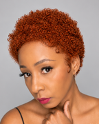 A close-up portrait of a woman with a short, tightly curled, textured wig in a warm coppery orange-brown shade, set against a solid gray studio background.