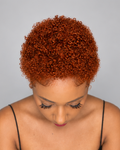 A woman with a short, tightly curled wig in a warm coppery orange-brown shade, looking down against a solid gray background.