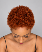 A woman with a short, tightly curled wig in a warm coppery orange-brown shade, looking down against a solid gray background.