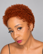 A woman with a short, tightly curled wig in a warm coppery orange-brown shade, looking down against a solid gray background.