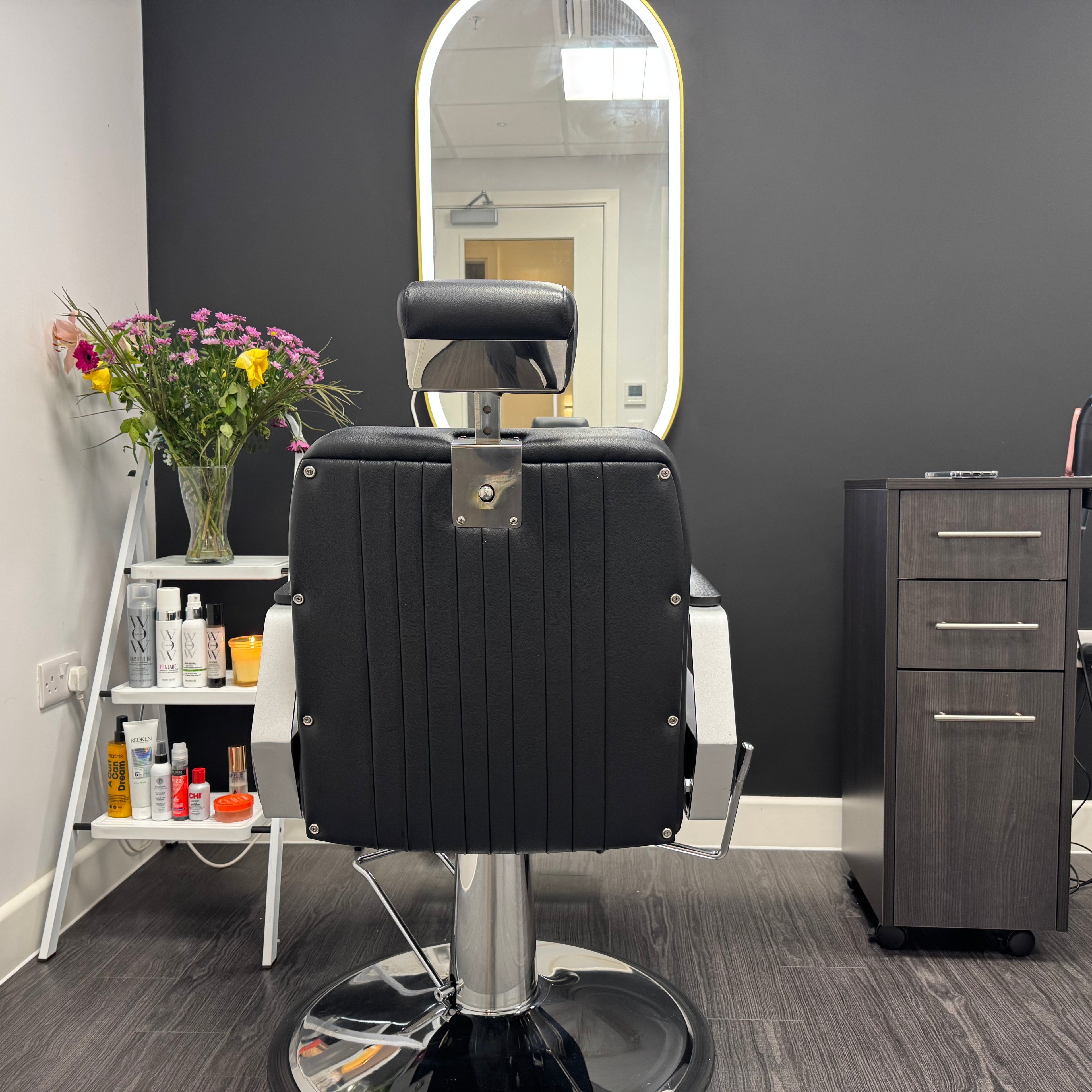 A black leather salon barber chair with chrome hardware, positioned in front of a large wall mirror in a modern hair salon. To the left is a white ladder shelf holding a vase of colorful flowers and various haircare products, and to the right is a dark wood rolling storage cabinet with a hair dryer on top.