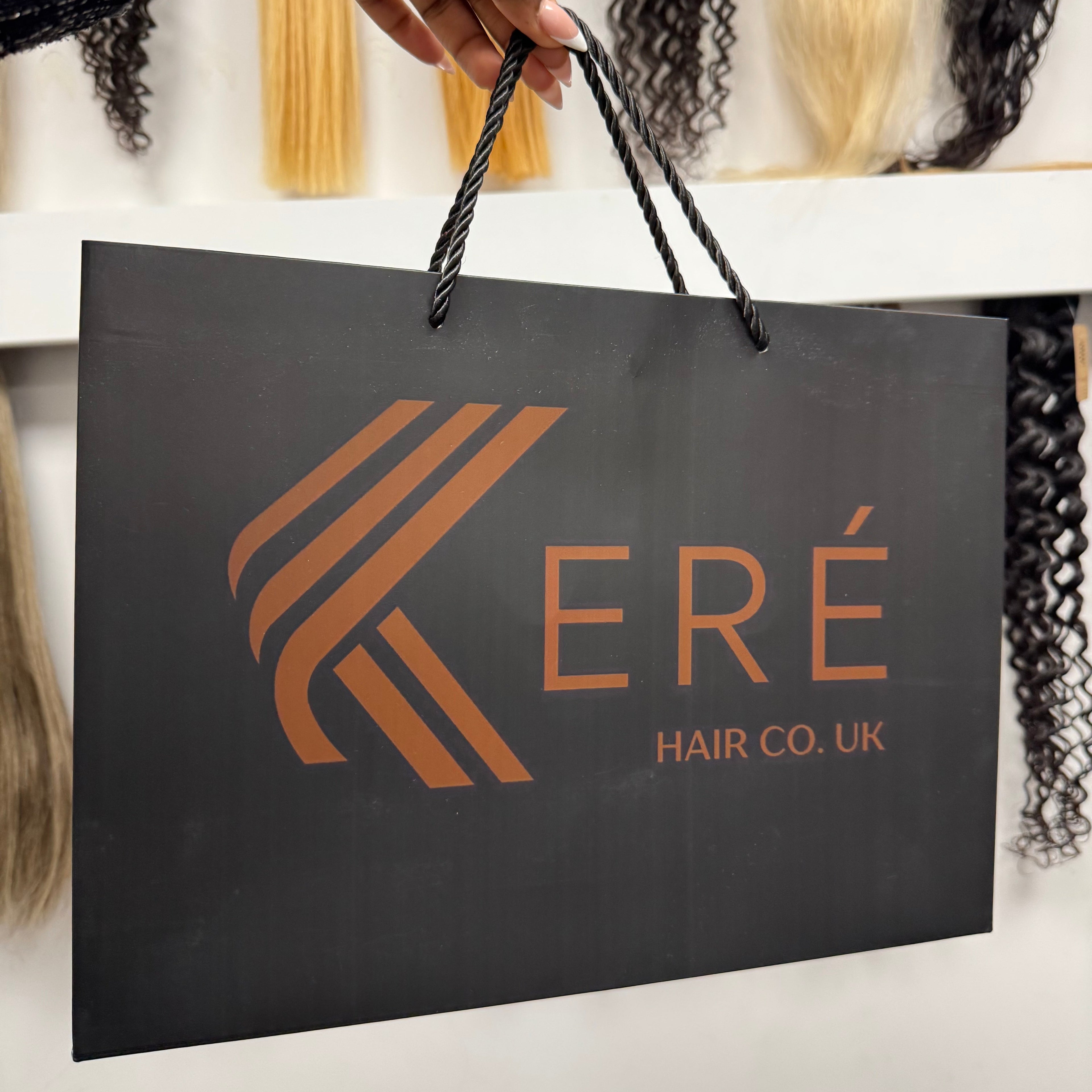 A hand holds a black paper shopping bag printed with the brand name KERÉ HAIR CO. UK' and a stylised logo in copper/bronze.
