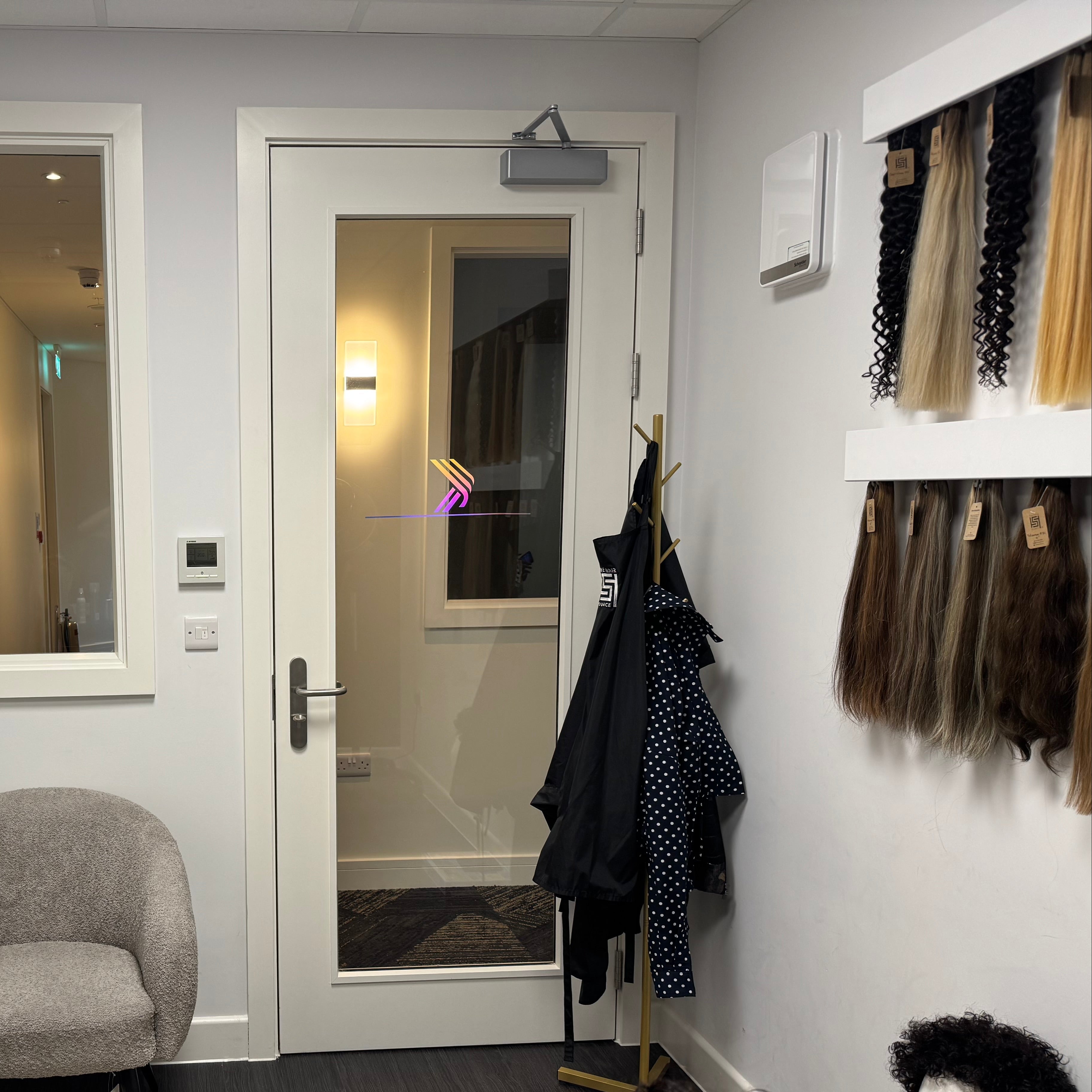 A hair color and styling consultation room at KERÉ HAIR CO. UK, featuring a gray armchair, a glass door, mannequin heads with wigs, and hair color swatches displayed on the right wall.