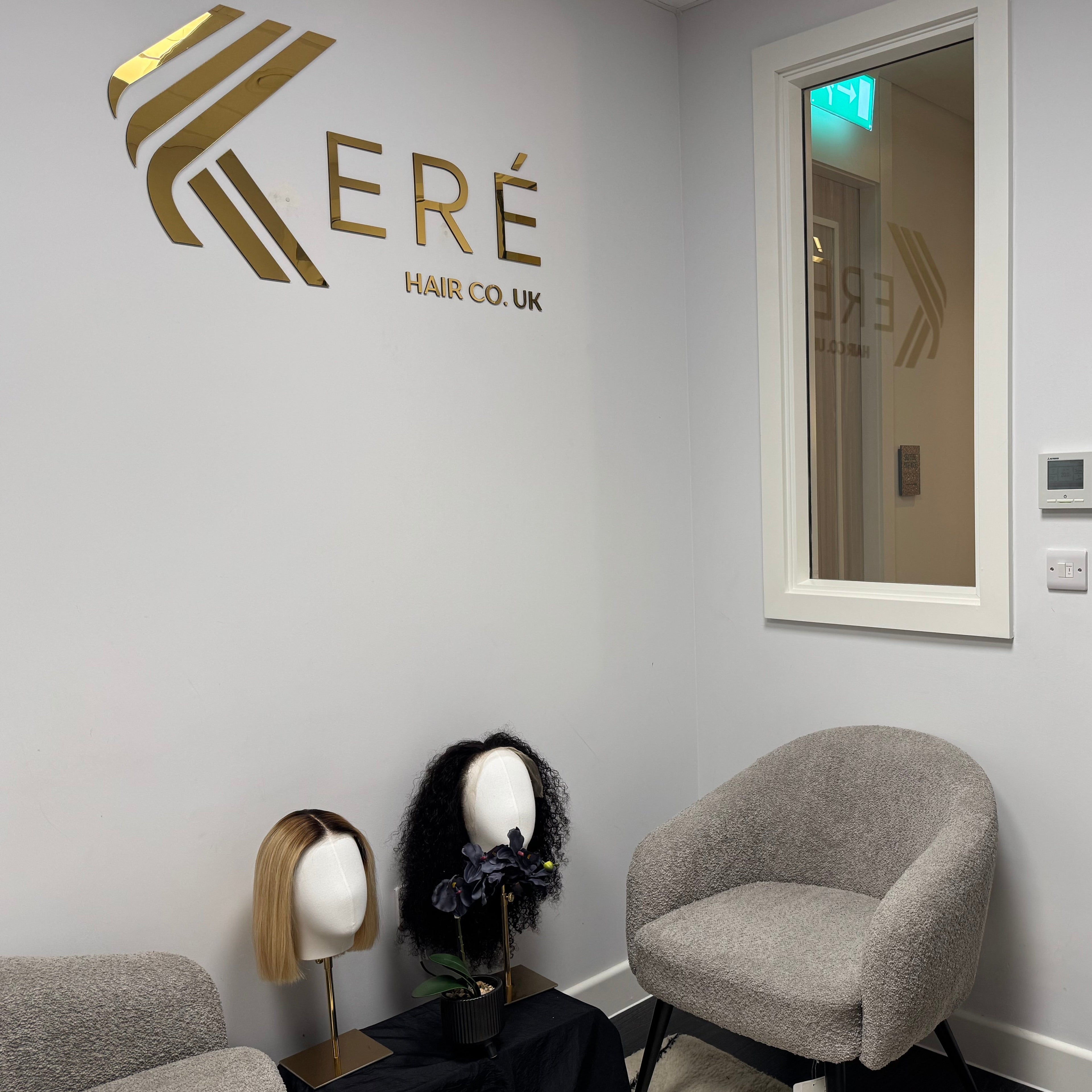 A hair salon waiting area with a gray upholstered armchair, a glass door, a mirror, and two mannequin heads displaying wigs on a side table. The wall features the gold Kéré Hair Co.