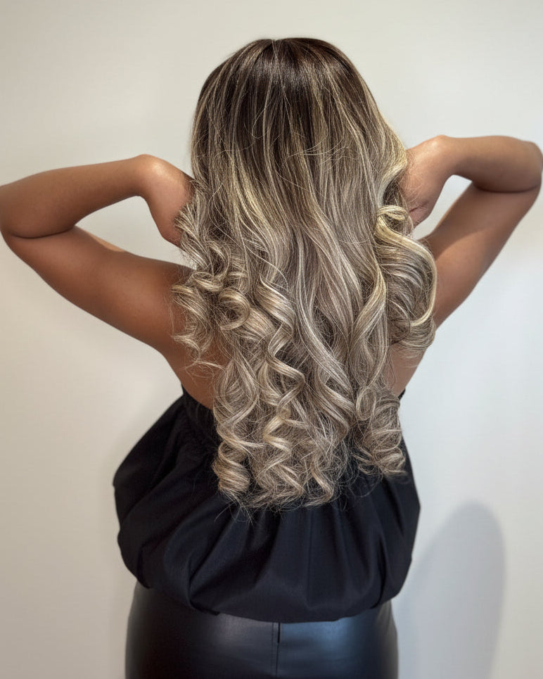 A woman shown from behind wearing a long, wavy wig with dark brown roots and blonde highlights, paired with a black dress against a plain light background.