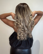 A woman shown from behind wearing a long, wavy wig with dark brown roots and blonde highlights, paired with a black dress against a plain light background.
