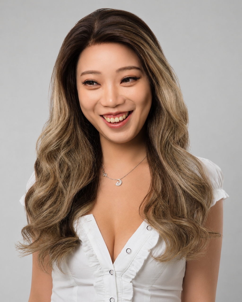A woman with long, wavy brown-to-blonde highlighted hair is shown from the chest up, wearing a white top against a plain light gray studio background.