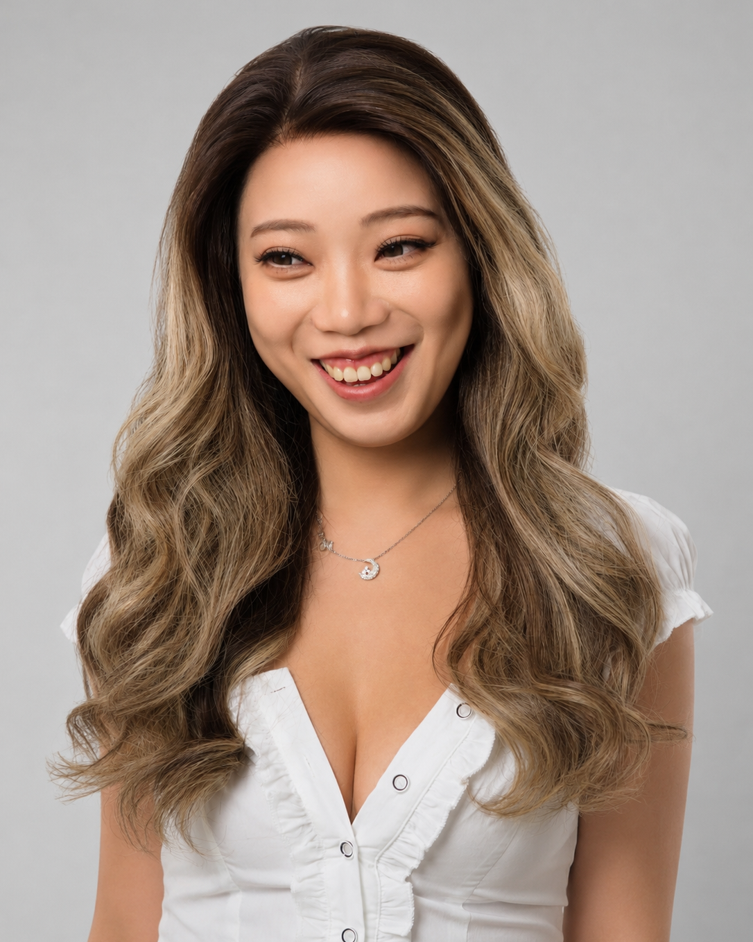 A woman with long, wavy brown-to-blonde highlighted hair is shown from the chest up, wearing a white top against a plain light gray studio background.