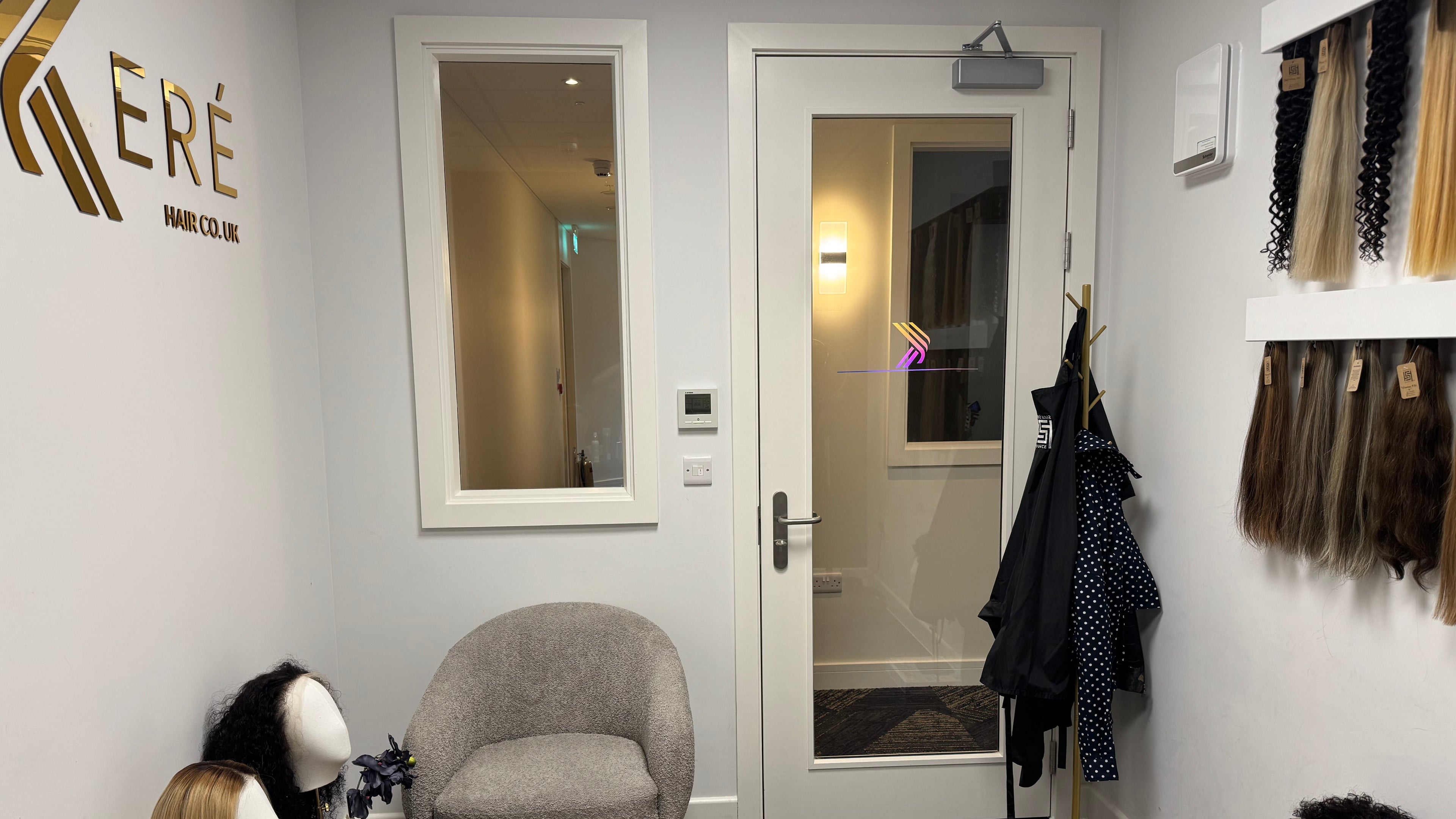 A hair styling consultation room at KERÉ HAIR CO. UK, featuring a grey armchair, a glass door, mannequin heads with wigs, and hair colour swatches displayed on the right wall.