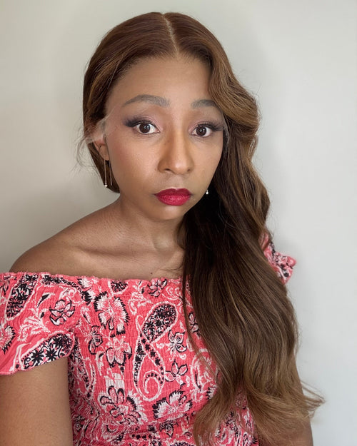 Woman with long brown hair wearing a pink and black patterned top against a plain background