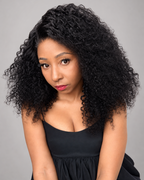 A woman with voluminous, tightly curled black hair wearing a black sleeveless top, photographed against a solid gray studio background.