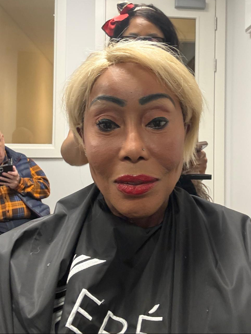 A person in a salon or beauty setting wearing a short blonde wig, a black salon cape, and makeup, with another person visible behind them and hair extensions hanging on the wall.