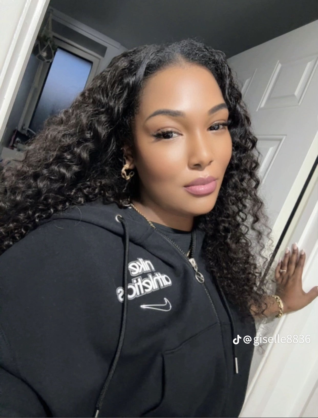 A woman wearing a black hoodie with visible branding, standing indoors, with Keré Haor Co UK Vietnamese raw hair extensions in a sew-in with leave-out style in a soft curly kink pattern.